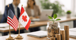 U.S. and Canadian flags displayed on a desk beside a glass jar filled with coins and a small green plant growing from the savings. Stacks of coins are arranged next to the jar, symbolizing cross-border investments, financial growth, and wealth building in the United States and Canada, with business professionals working in the background.